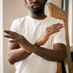 Crop unrecognizable bearded African American male in white t shirt demonstrating no gesture with hands while standing in light room